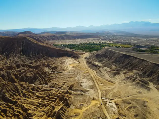 Vista aérea de un camino de tierra entre quebradas del desierto que baja hacia un oasis verde con cordillera al fondo