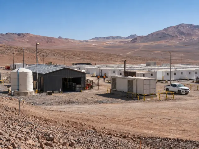 Campamento de módulos blancos en hileras en el desierto, con bodega metálica, estanque de agua, postes de luz y un grupo electrógeno en contenedor